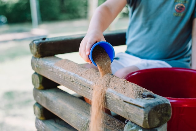 Eine Kinderhand schüttet Sand aus einem Becher auf ein Holzgeländer.